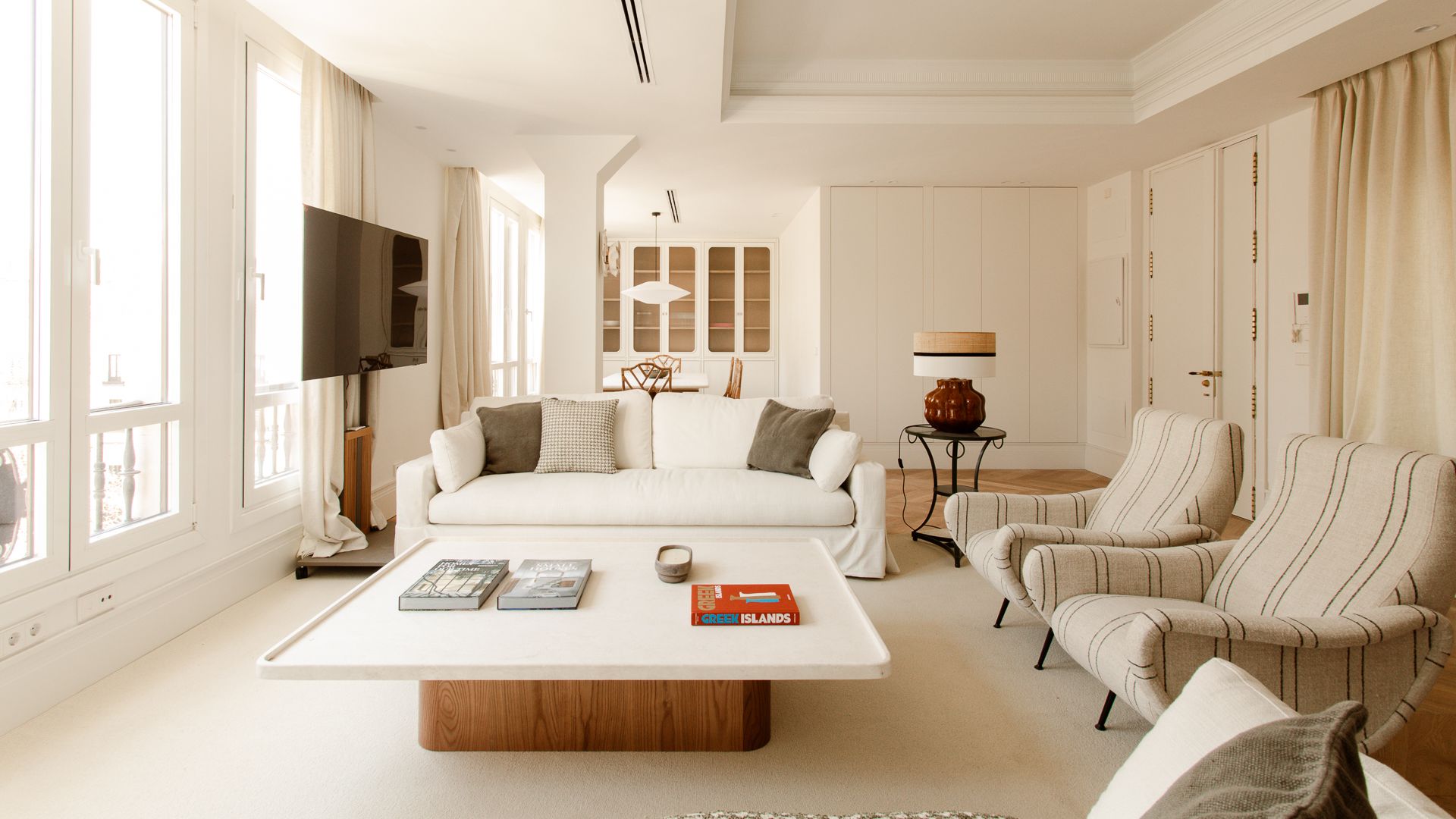 Salón comedor blanco con grandes ventanales, sofá blanco, alfombra, mesa de centro blanca y de madera, butacas tapizadas en tela blanca con rayas, mueble de televisión