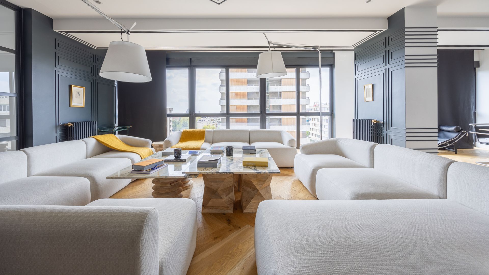 Salón con sofás blancos XL, techo decorado, pared con molduras de color azul, radiadores de hierro a juego, suelo de madera, mantas amarillas en el sofá, mesa de centro y dos lámparas de pie blancas 