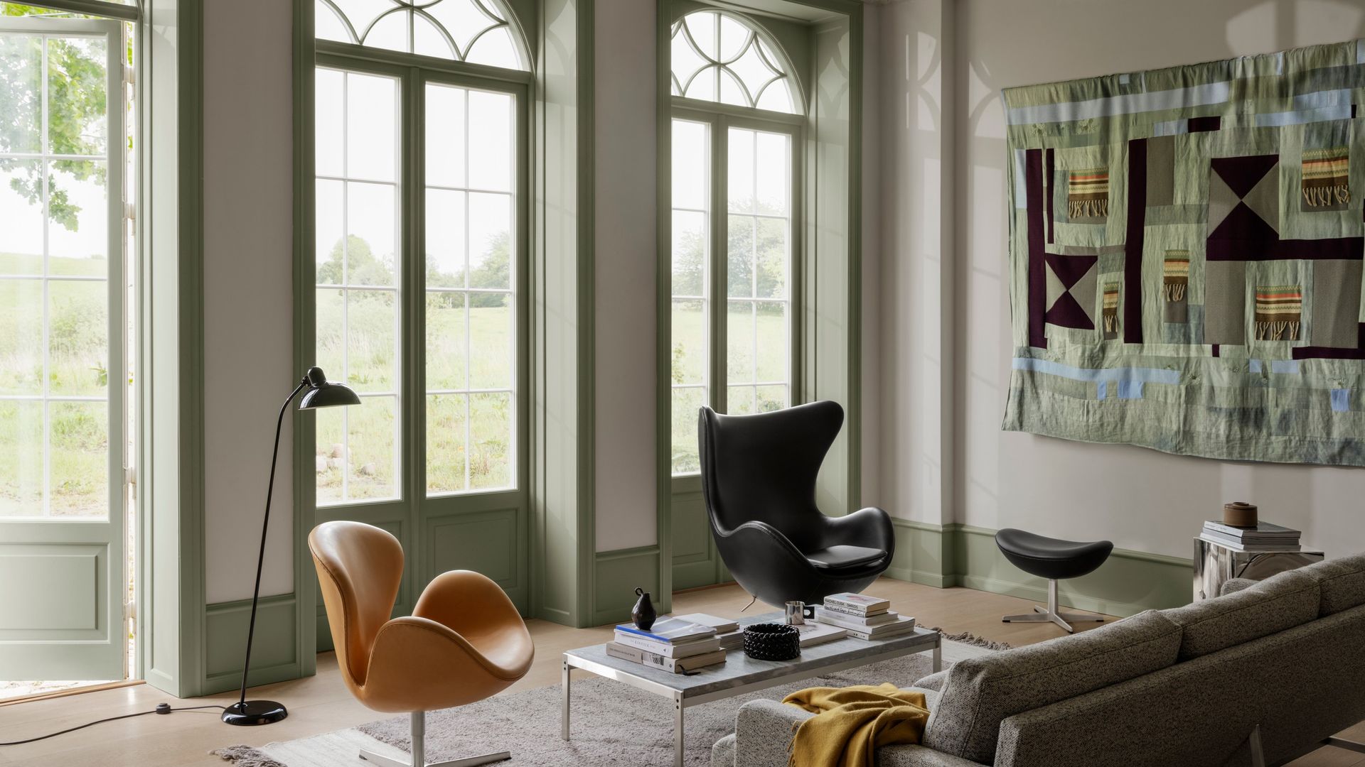 Salón con sofá gris, alfombra, butacas de cuero marrón y negro, lámpara de pie, ventanas y puertas de color verde claro y tapiz en la pared