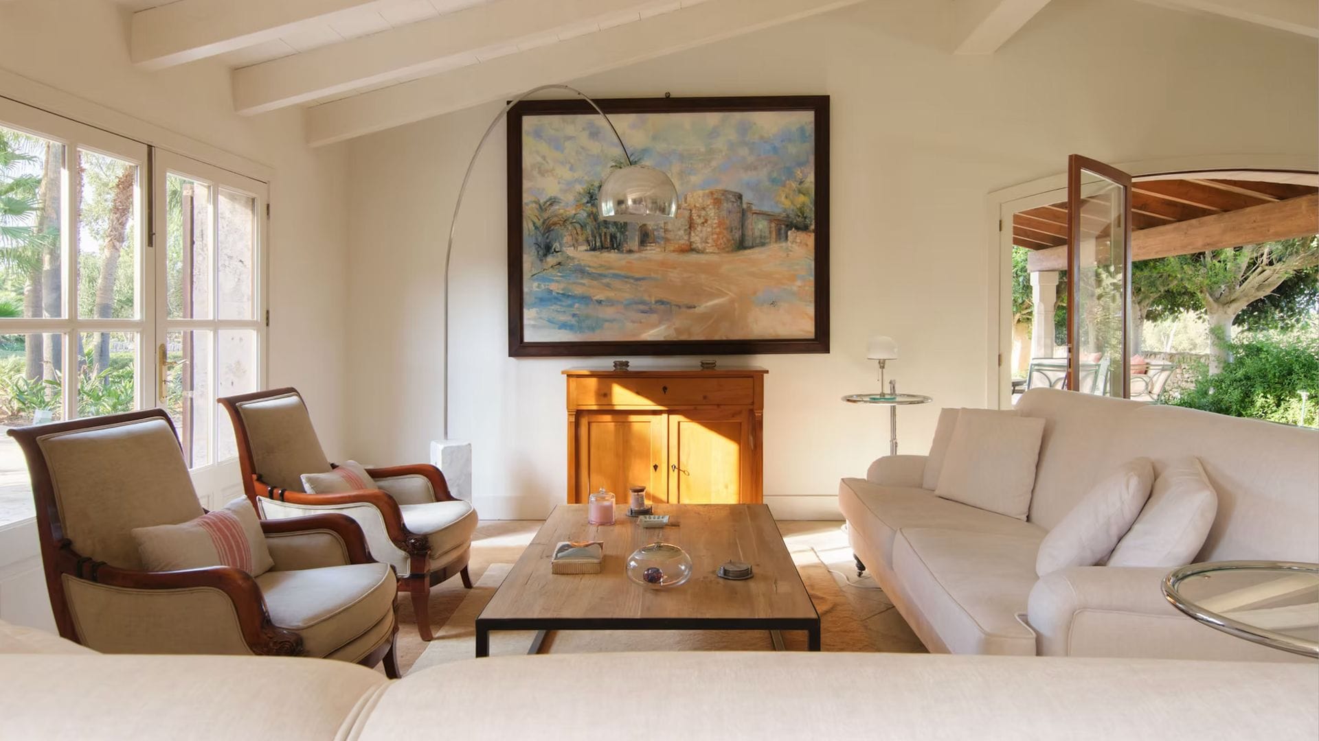 Salón con vigas de madera blanca en el techo, suelo de madera, aparador de madera, cuadro, mesa de centro de madera, sofá blanco, lámpara de pie tipo Arco