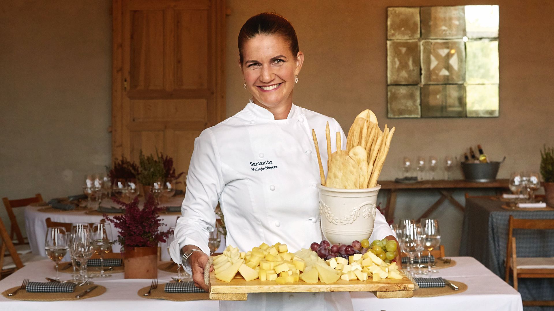 Samantha Vallejo-Nágera, en uniforme de chef, sostiene una tabla con queso y uvas.