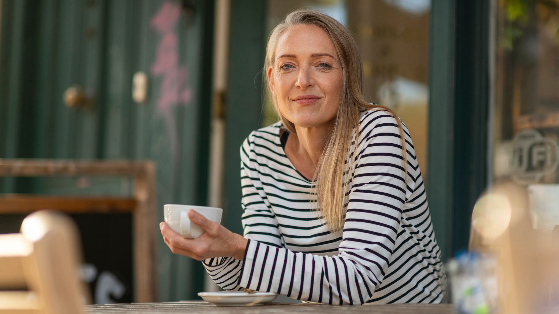 Mujer de 50 años con una taza de café entre manos, sentada en un mesa de madera de la terraza de una cafetería