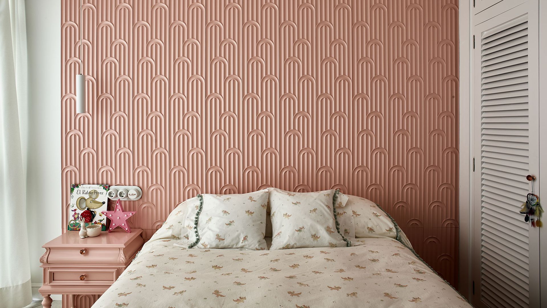 Habitación infantil con papel pintado rosa, mesita de noche rosa, armario de lamas blancas, ropa de cama blanca con pequeños motivos