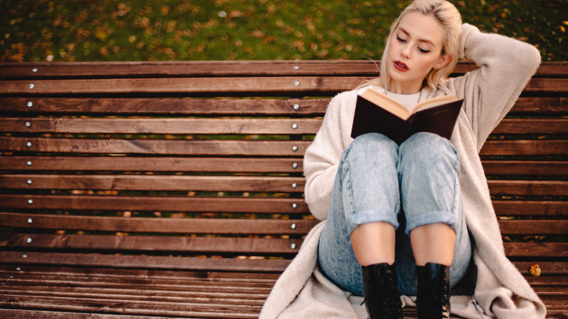 Chica joven rubia leyendo en un parque, sentada en un banco de madera, con ropa de otoño. Lleva vaqueros, unas botas Dr. Martens y una rebeca 'beige'.