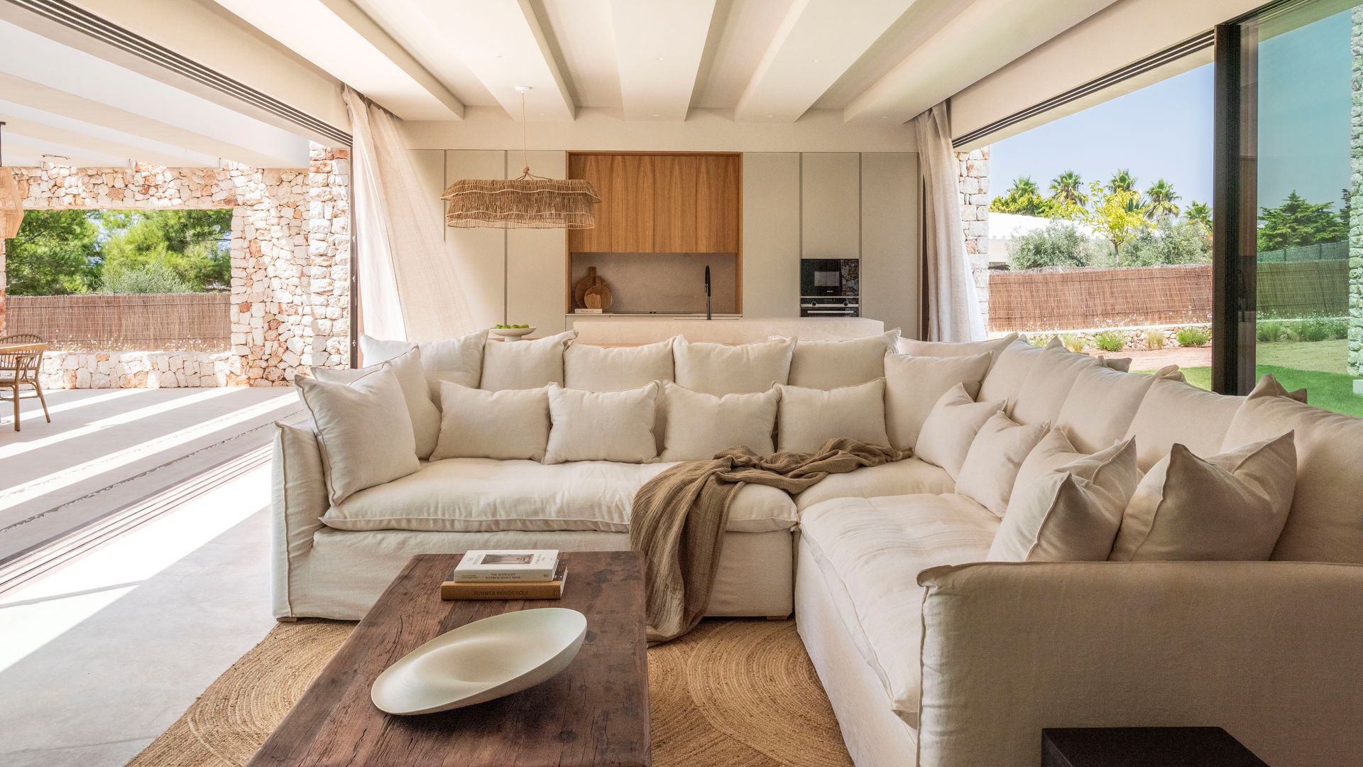 Salón de estilo mediterráneo con cocina, ventanales a ambos lados con salida al jardín, sofá beige, lámpara de techo de fibras vegetales