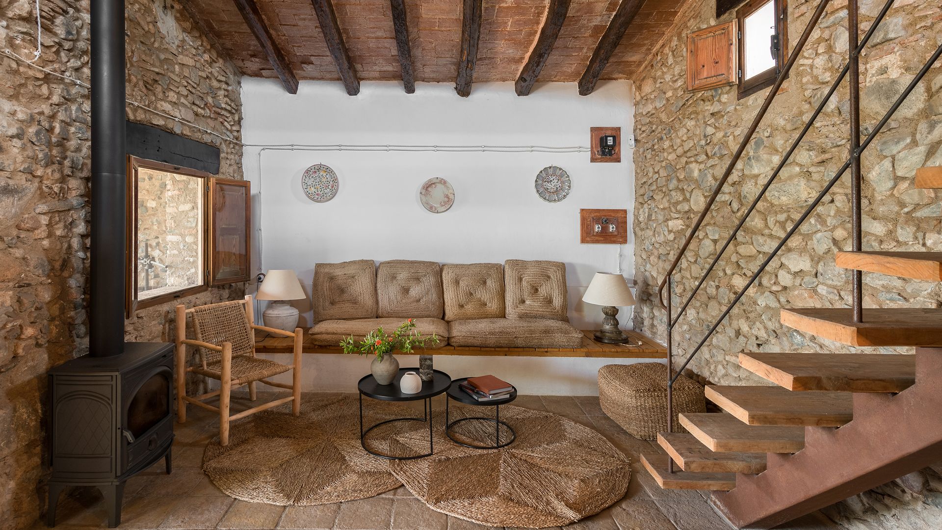 Salón casa de campo con sofá de obra, alfombra redonda de fibras vegetales, platos en la pared del sofá, bóvedas catalanas en el techo 