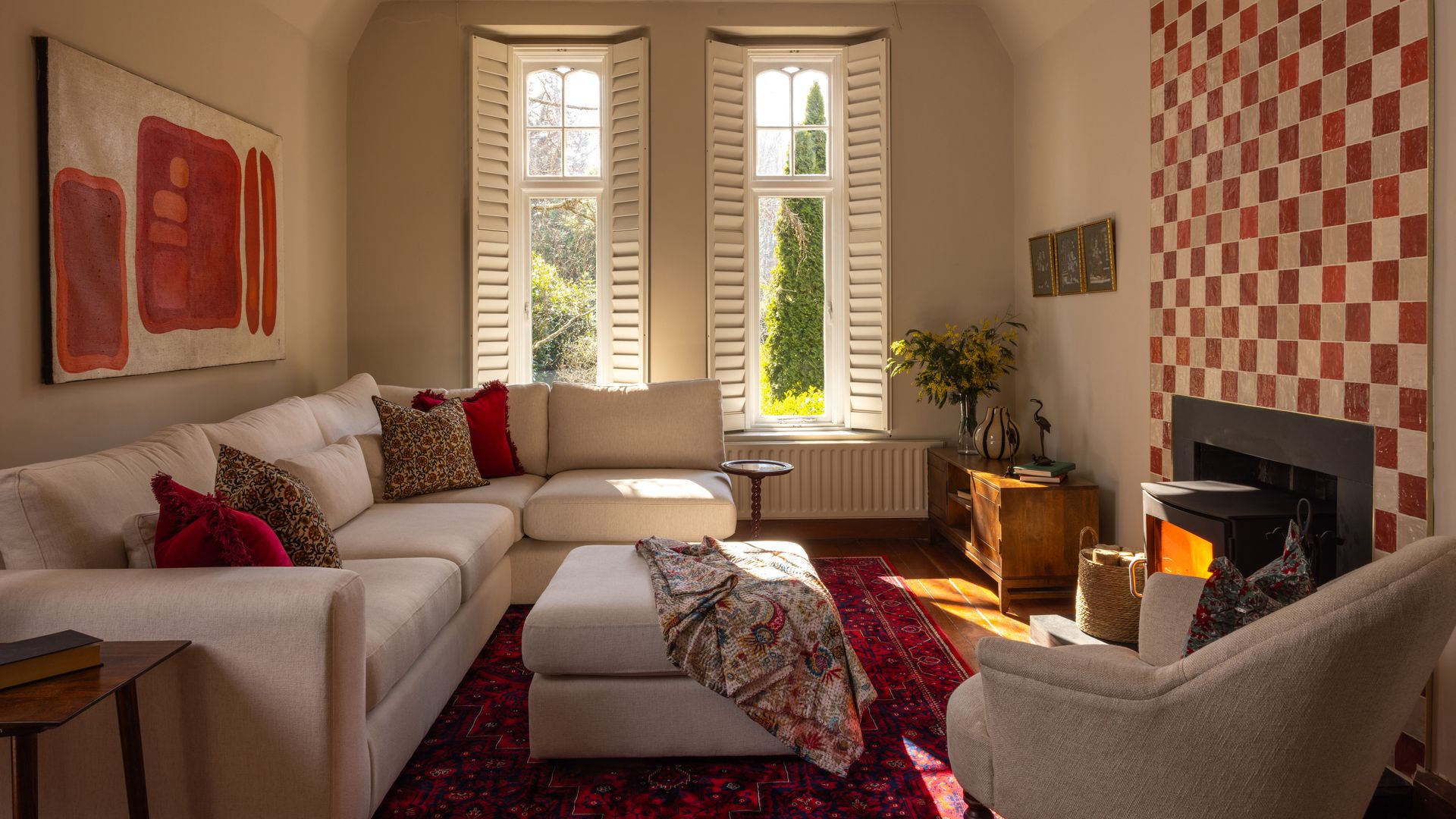 Salón con chimenea, pared de azulejos rojos y blancos, alfombra roja, sofá blanco, mesa de centro tapizada, cuadro en la pared del sofá, contraventanas blancas, butaca