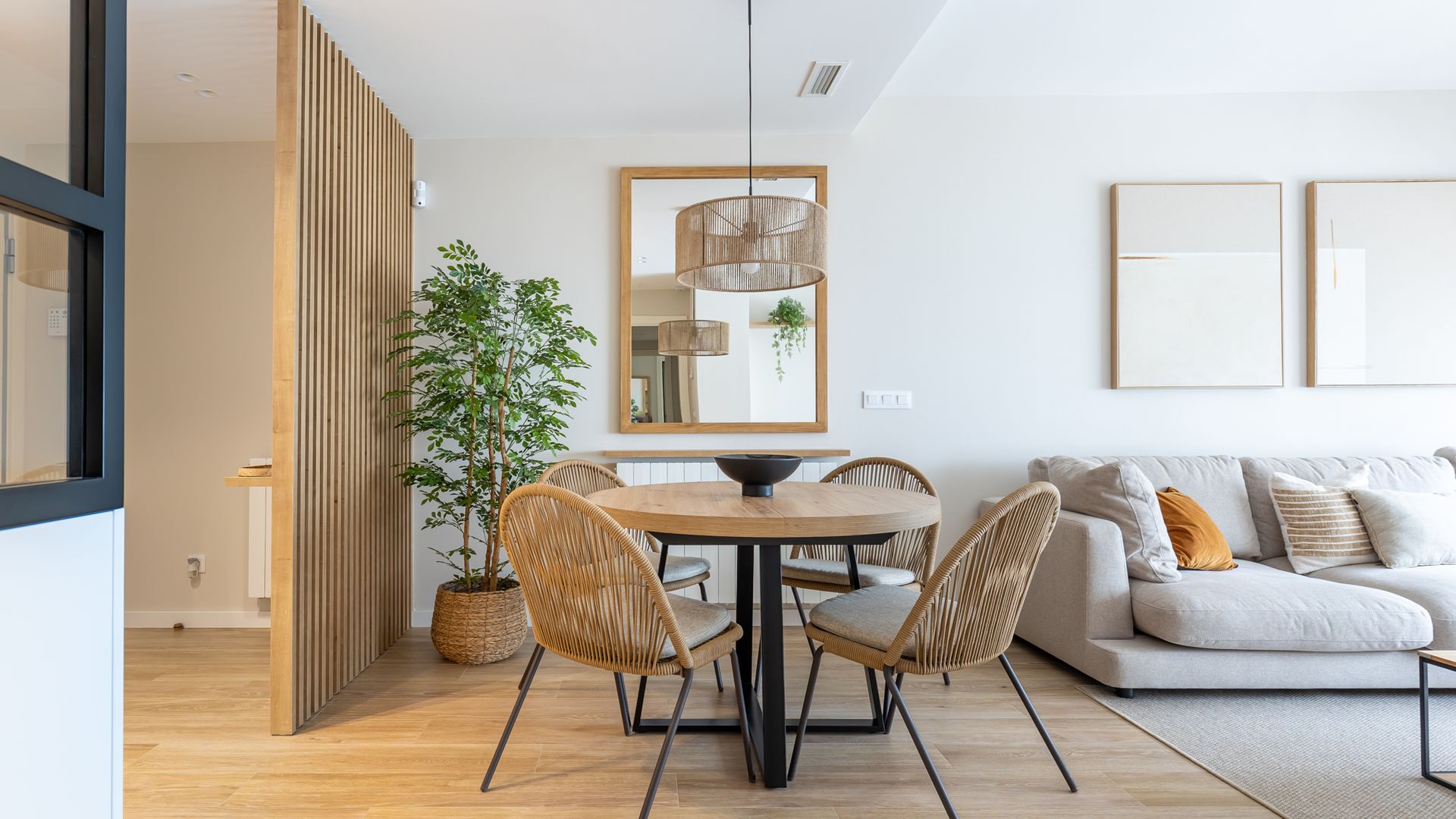 Salón comedor con mesa redonda de madera y pie negro, sofá gris, alfombra gris, espejo con el marco de madera, sillas de fibras vegetales, lámpara de techo de fibras vegetales, tabique de lamas de madera, separando el recibidor y tabique de cristal con cuarterones negros separando la cocina