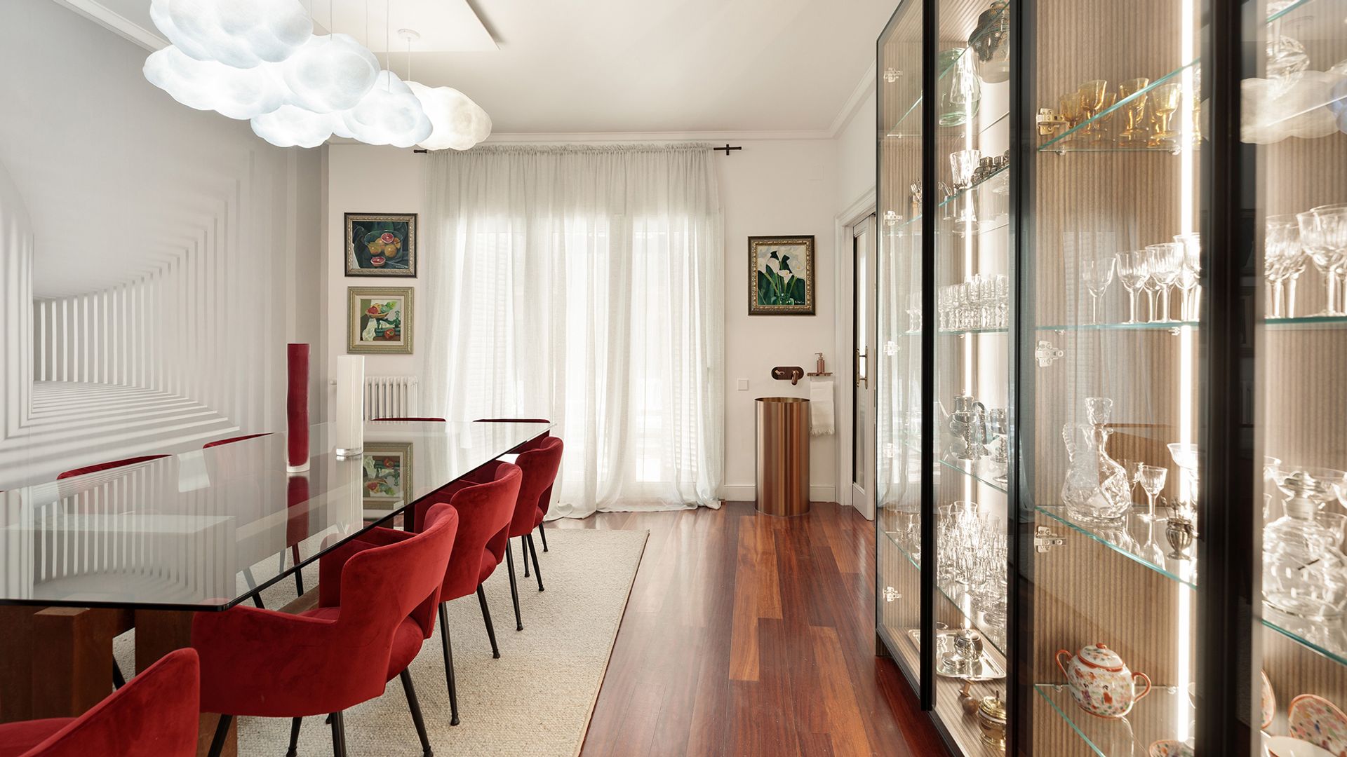 Comedor con gran mesa de cristal, suelo de madera oscuro, vitrina doble con vajilla y cristalería, lámparas de techo blancas, cortinas blancas, alfombra, fuente, cuadros en la pared, sillas tapizadas en rojo