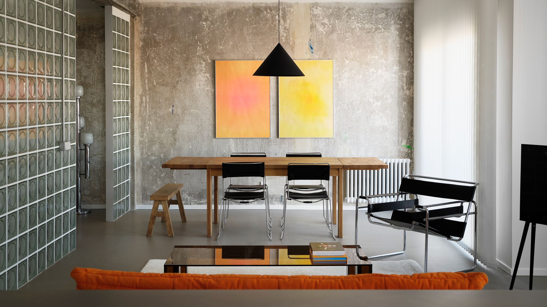 Comedor con mesa de madera, pared de hormigón, tabique de payés, cuadros en la pared, sillas metálicas con el respaldo y el asiento negros, sillón Wassily y lámpara de techo negra
