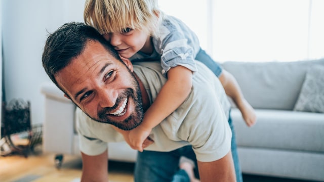 Padre jugando con su hijo a la espalda en el salón de su casa, sonriendo.