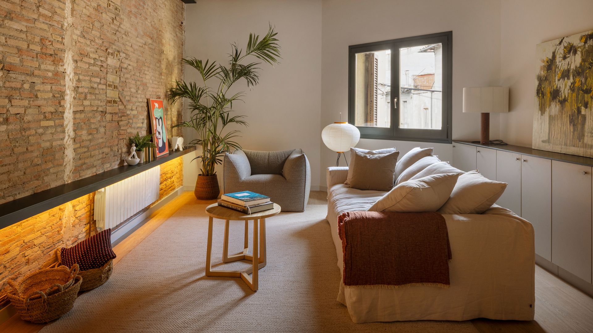 Salón con radiador, pared de ladrillo, mueble bajo la ventana, vigas en el techo, planta en el suelo, alfombra