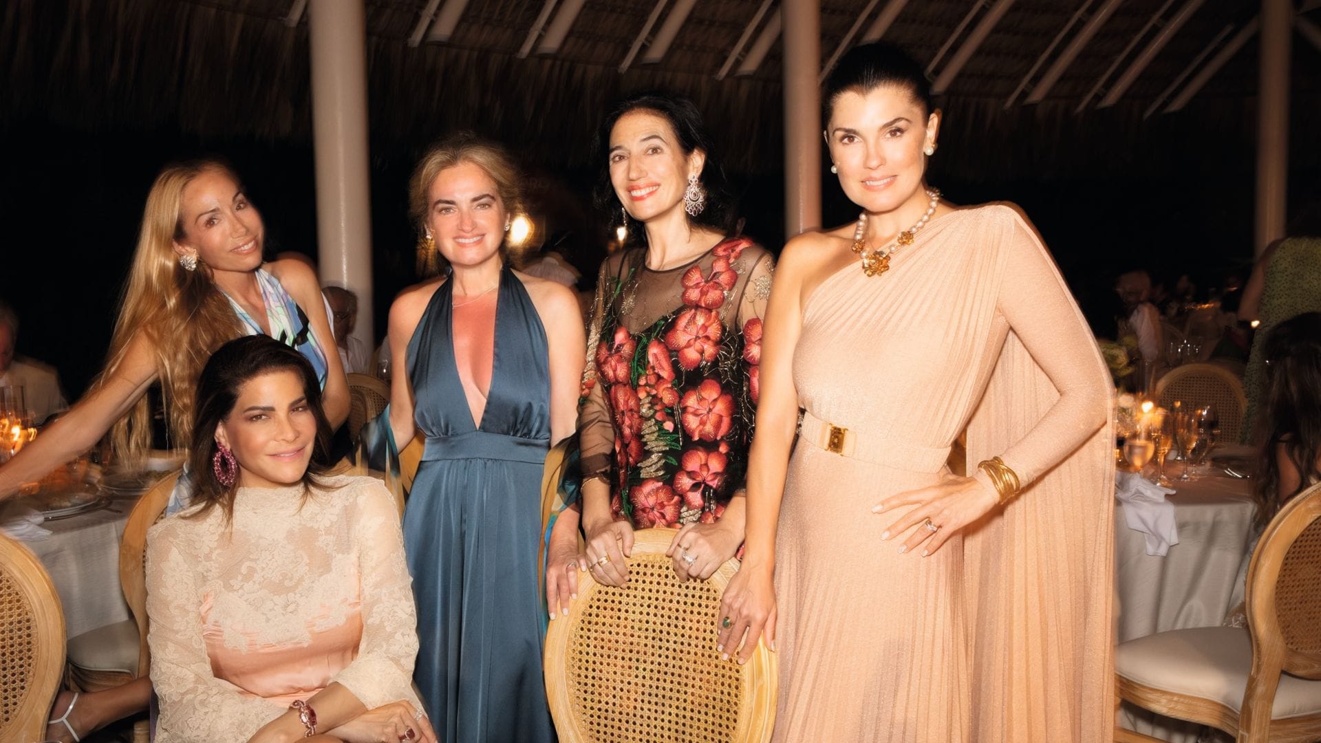 Cinco mujeres en una cena elegante, con vestidos variados y joyas, posando juntas.