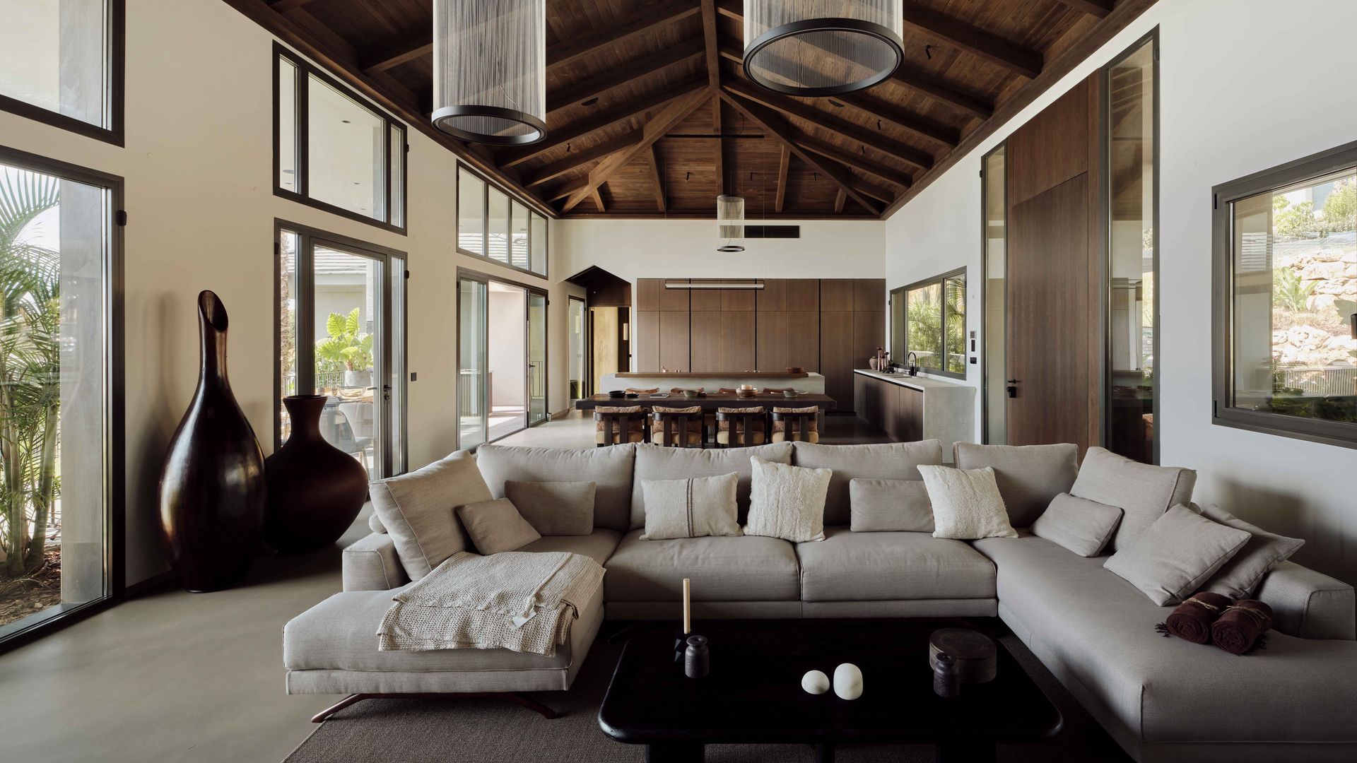 Salón comedor con sofá beige XL, mesa negra, techo de madera, alfombra, ventanales, lámparas de techo XL, vasijas muy grandes