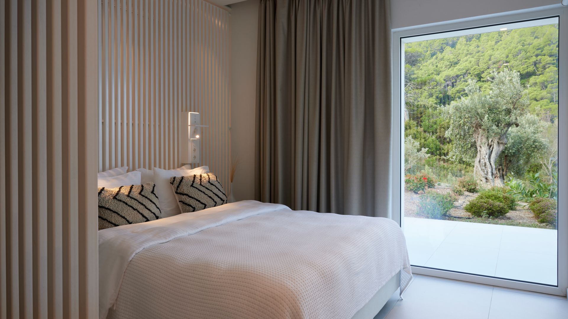 Dormitorio principal con pared del cabecero de lamas de madera, cortinas beige oscuro, ropa de cama beige, cojines marrones sobre la cama, ventanal con salida al jardín 
