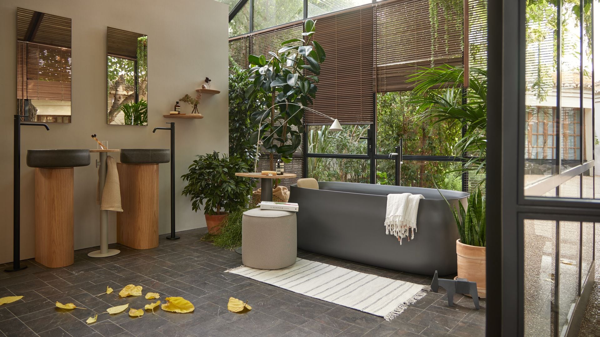 Baño con bañera exenta gris, techo y paredes de cristal, lavabos de pedestal, espejos, plantas, lámpara de pie, alfombra, estores de madera,