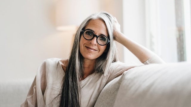 Mujer sonriendo a la cámara, sentada en un sofá en tonos claros. Tiene el pelo muy largo y entre blanco y gris, con gafas y sonriendo.