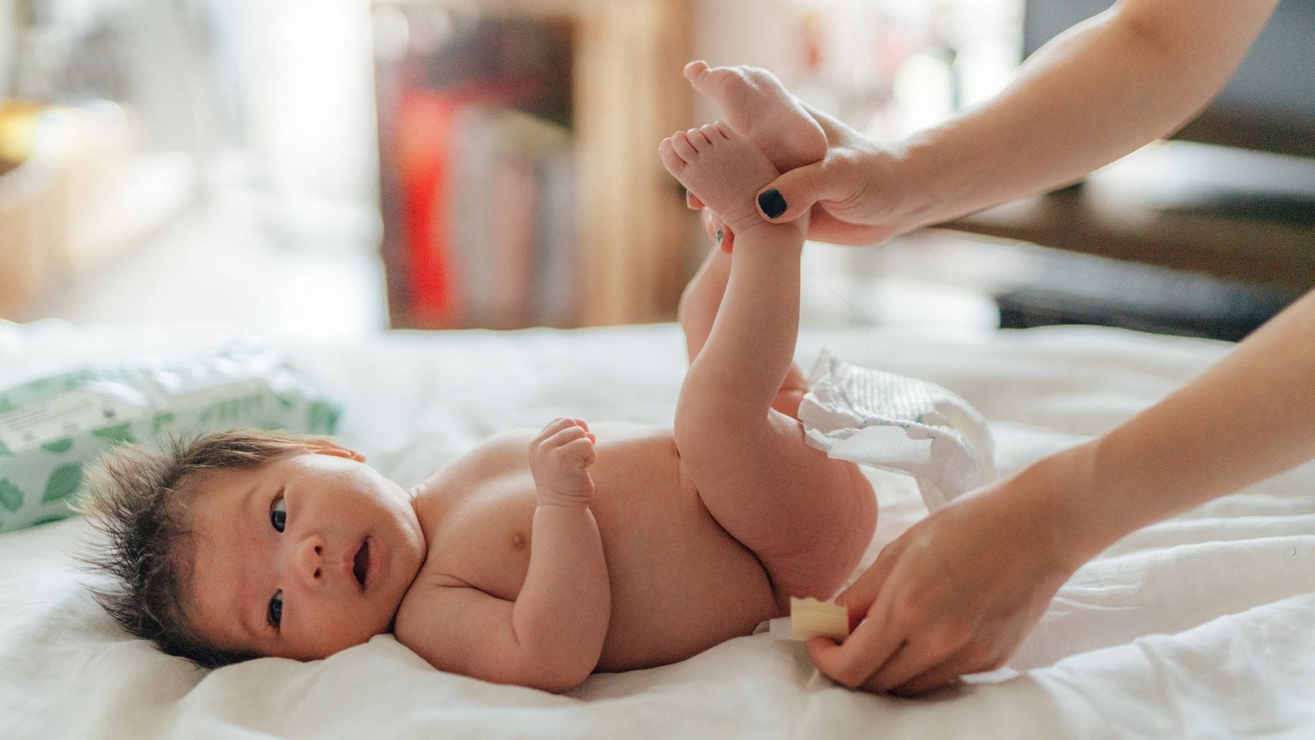 Madre cambiando el pañal de su bebé encima de la cama. El bebé está sin ropa mirando hacia un lado. 