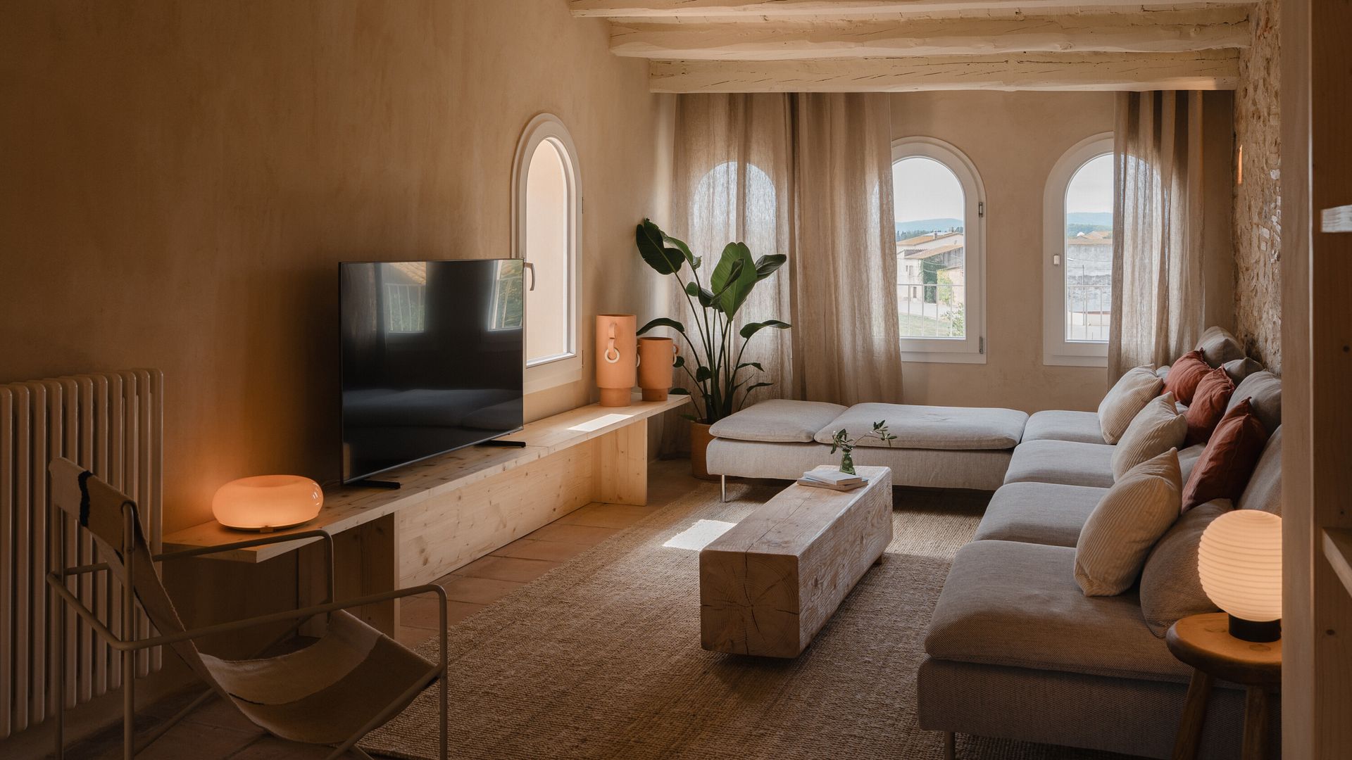 Salón con mueble de televisión de madera, televisión, sofás con chaise longue, alfombra, vigas de madera, cortinas 