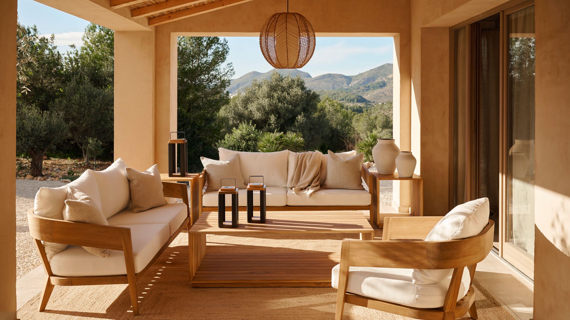 Porche con salón exterior con sofás y sillones de madera, asientos y respaldos blancos, alfombra y lámpara de techo de fibras vegetales