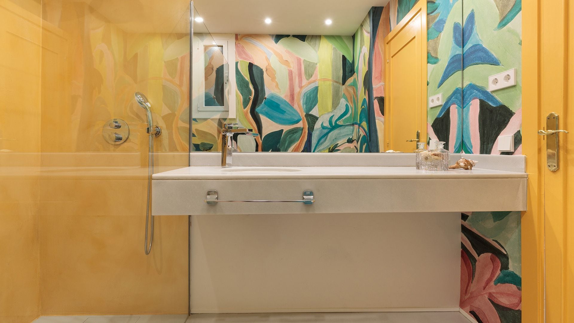 Baño con ducha, encimera volada, puerta amarilla, papel pintado de colores y motivos tropicales, mampara 
