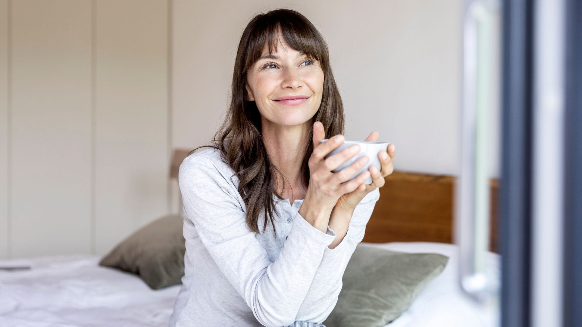 Mujer morena sonriente sentada en su cama con una taza de café