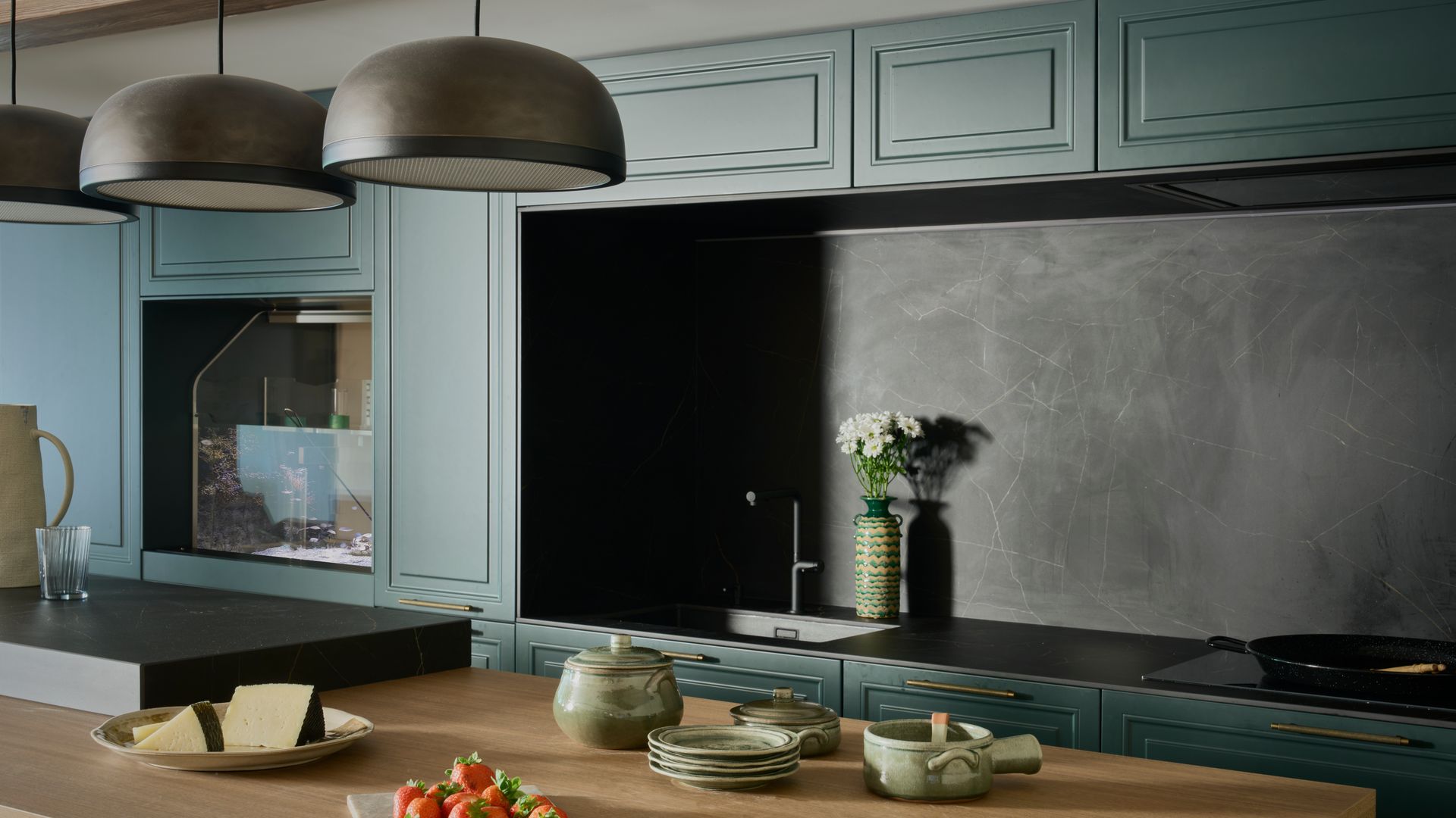 Cocina con mesa de cocina de madera, isla, muebles verde oscuro, lámparas de techo negro sobre la mesa, antepecho de cocina negro