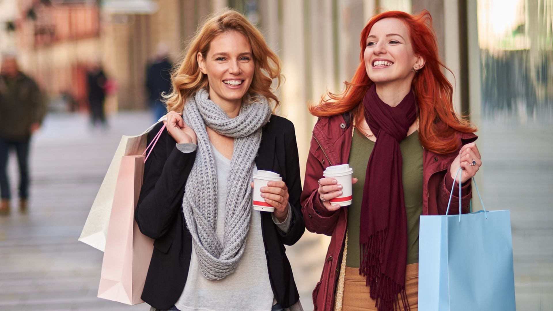 Dos mujeres paseando por la calle con bolsas de shopping y dos cafés para llevar en la mano. Van sonrientes con ropa de otoño-invierno.