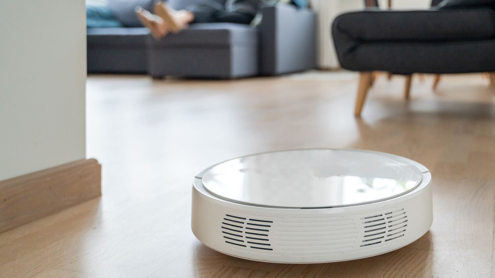 Mujer relajada en el sofá de su casa (desenfocada) mientras un robot aspirador (en primer plano) limpia el suelo de la casa.