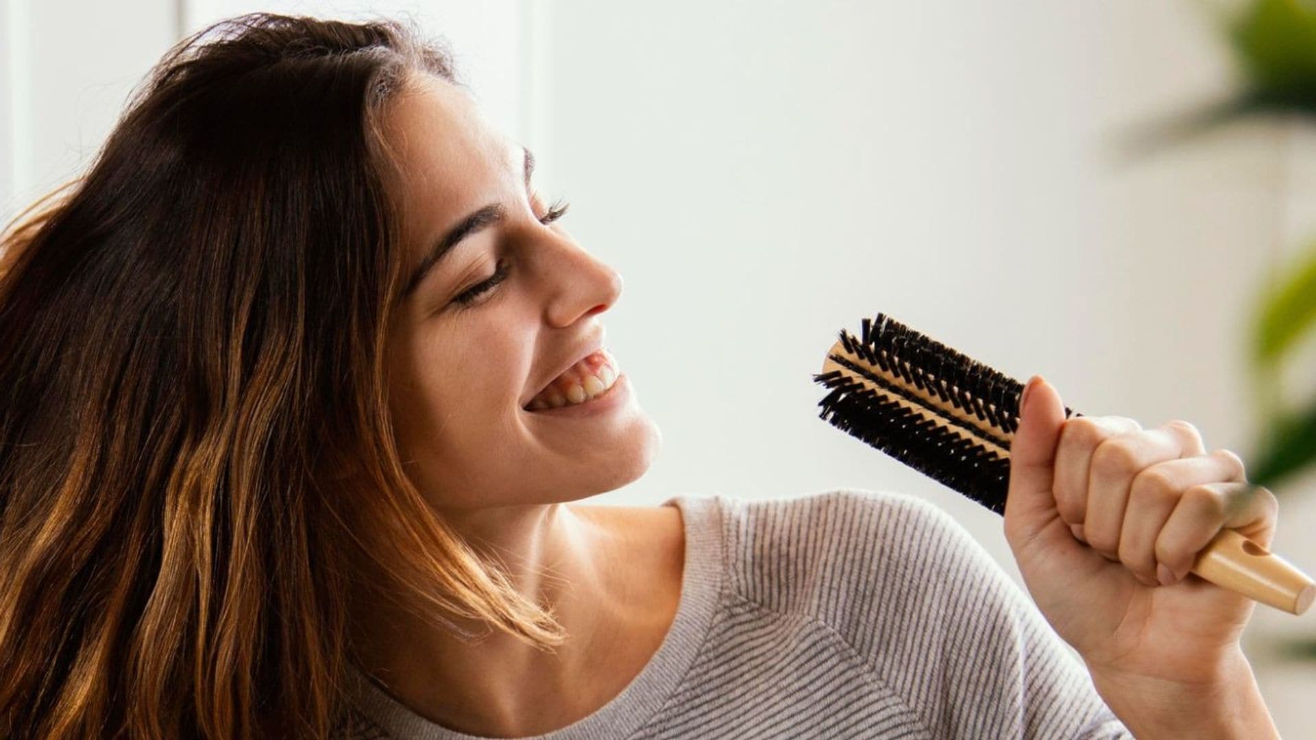 Mujer cantando con un cepillo de cabello, sonriente. Tiene melena larga y castaña.