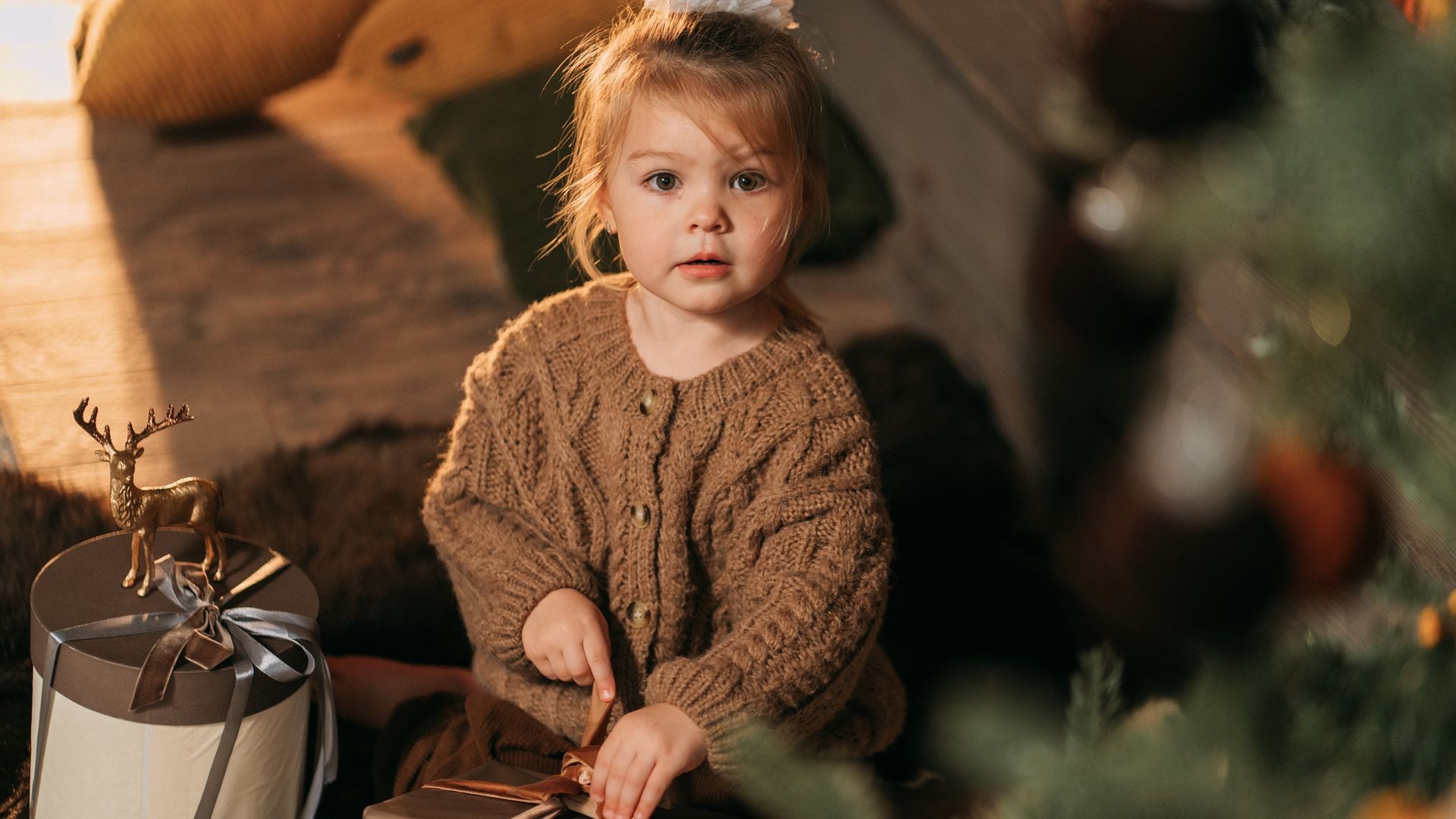 Niña de tres o cuatro años abriendo un regalo de Navidad al lado del árbol. Está mirando hacia la cámara y viste en tonos marrones.