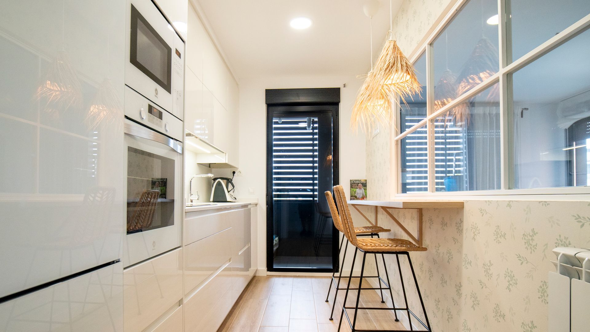 Cocina blanca con barra de desayunos, tabique de cristal que comunica con el salón, lámparas de fibras, taburetes, suelo de madera