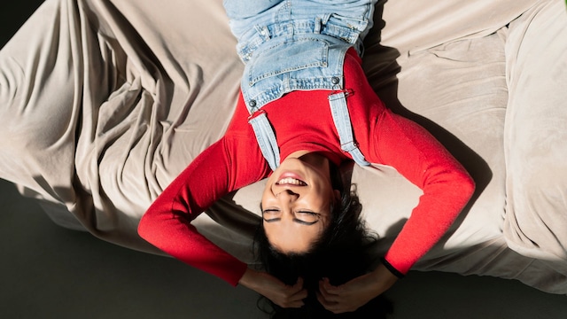 Mujer joven tumbada sobre un sofá sonriendo y feliz, con un jersey rojo y un peto vaquero, dándole la luz en la cara
