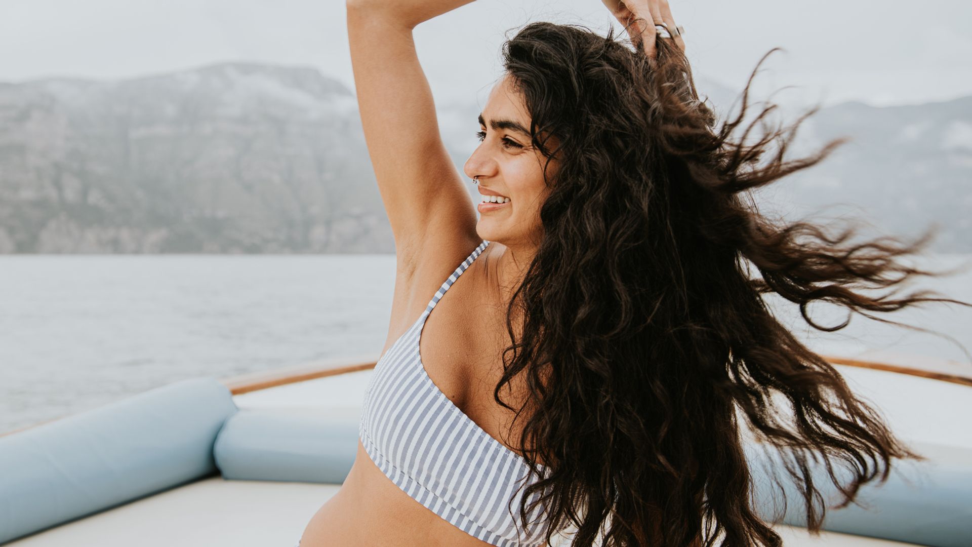 Mujer joven con bikini de rayas en un barco, tocándose la melena negra y larga con la mano