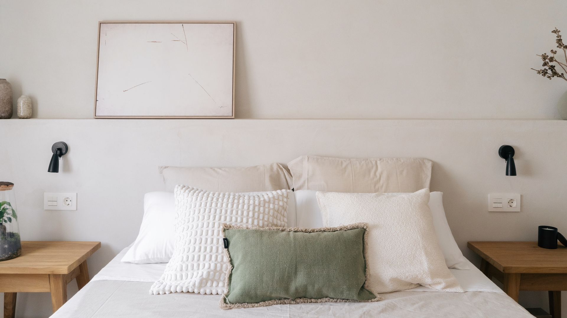 Dormitorio con cabecero de obra con repisa, cuadro en la repisa, mesitas de noche de madera, apliques de pared negros, ropa de cama beige y cojines