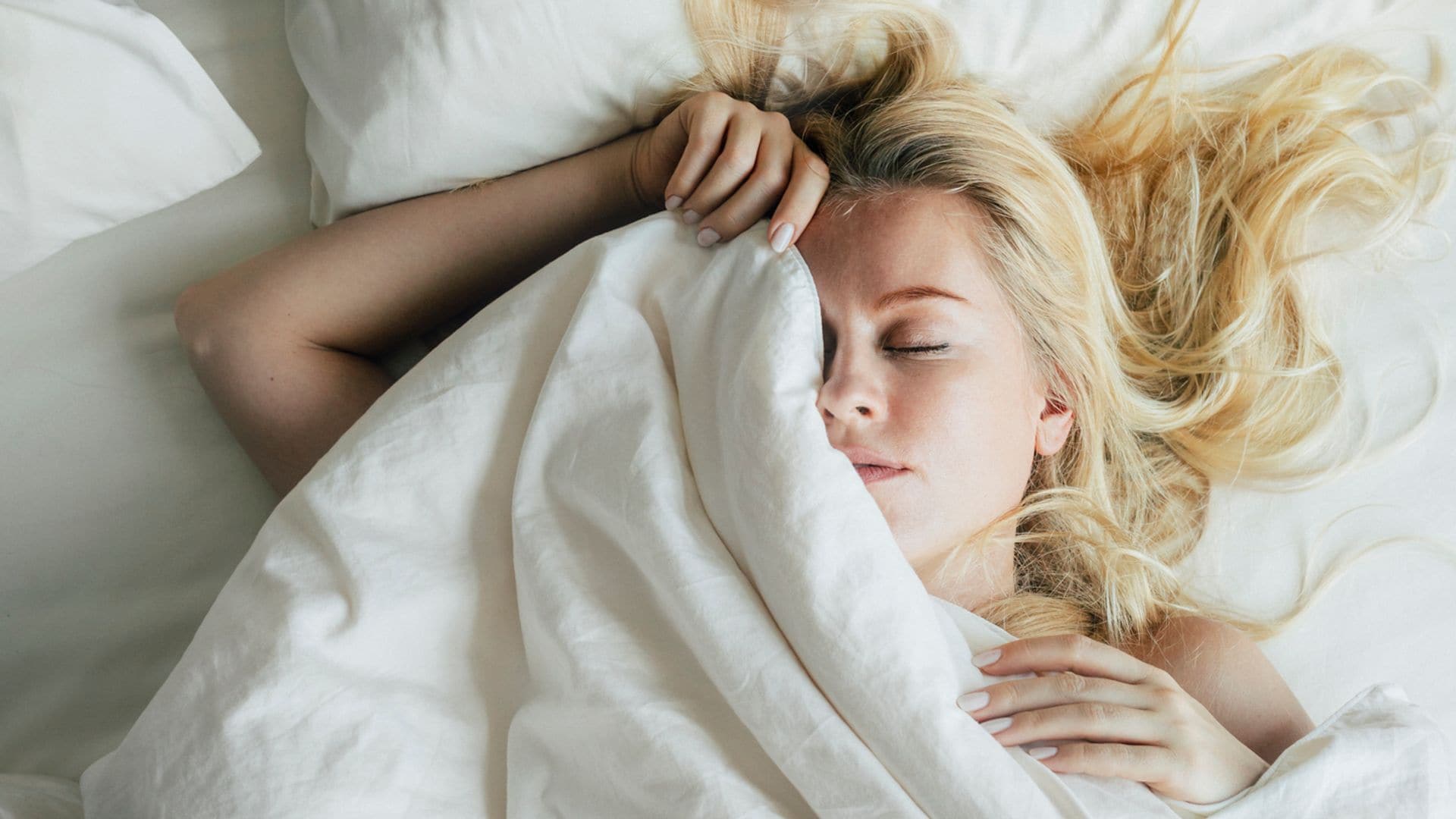 Mujer joven rubia tumbada en la cama blanca, tapándose con el edredón la mitad de su cara. El plano es cenital desde arriba.