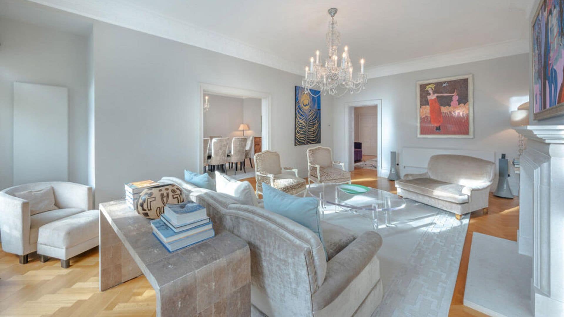 Salón con chimenea, lámpara de araña, alfombra, sofás y butacas de terciopelo beige, mesa de centro transparente 