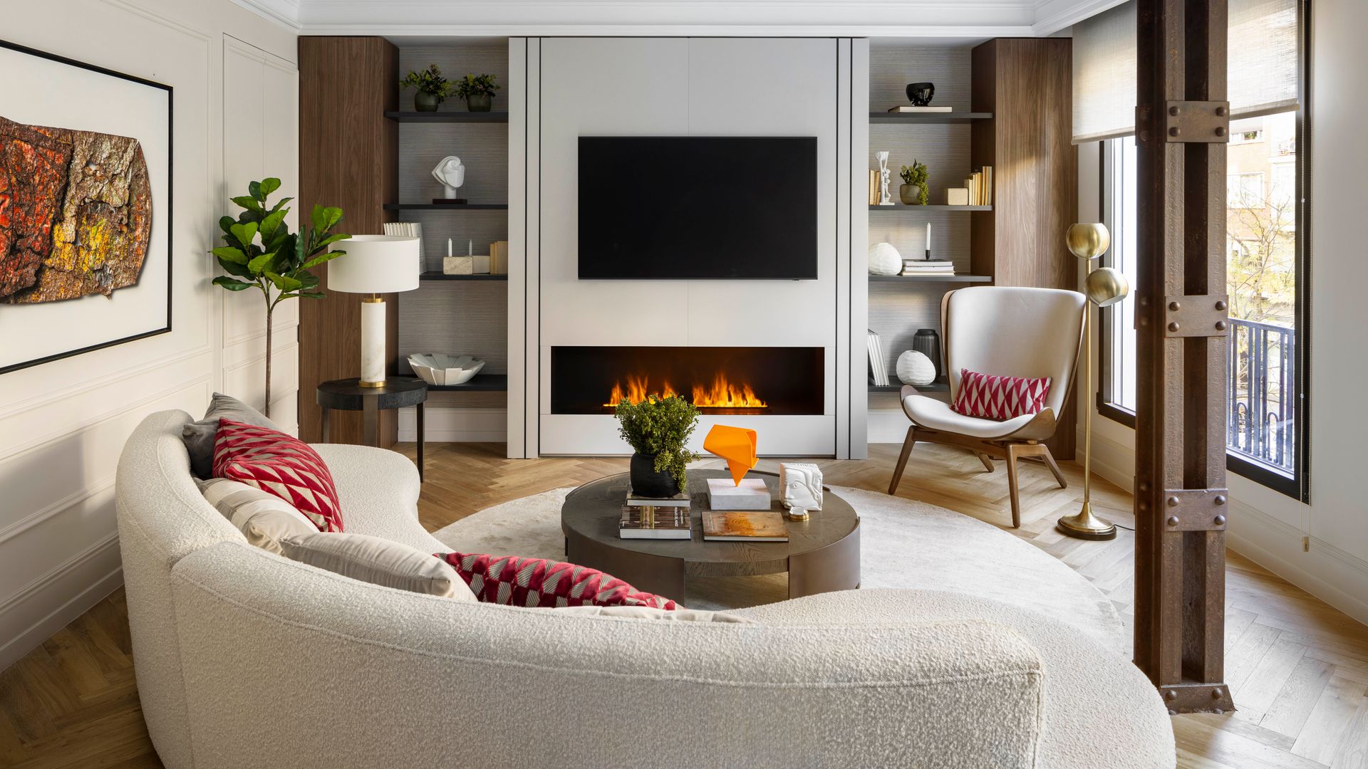 Salón con chimenea, viga metálica, televisión colgada con estanterías a los lados, sofá blanco de lana bouclé, cojines rojos, alfombra