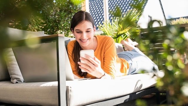 mujer tumbada al sol en un sofá en su terraza, mirando su móvil