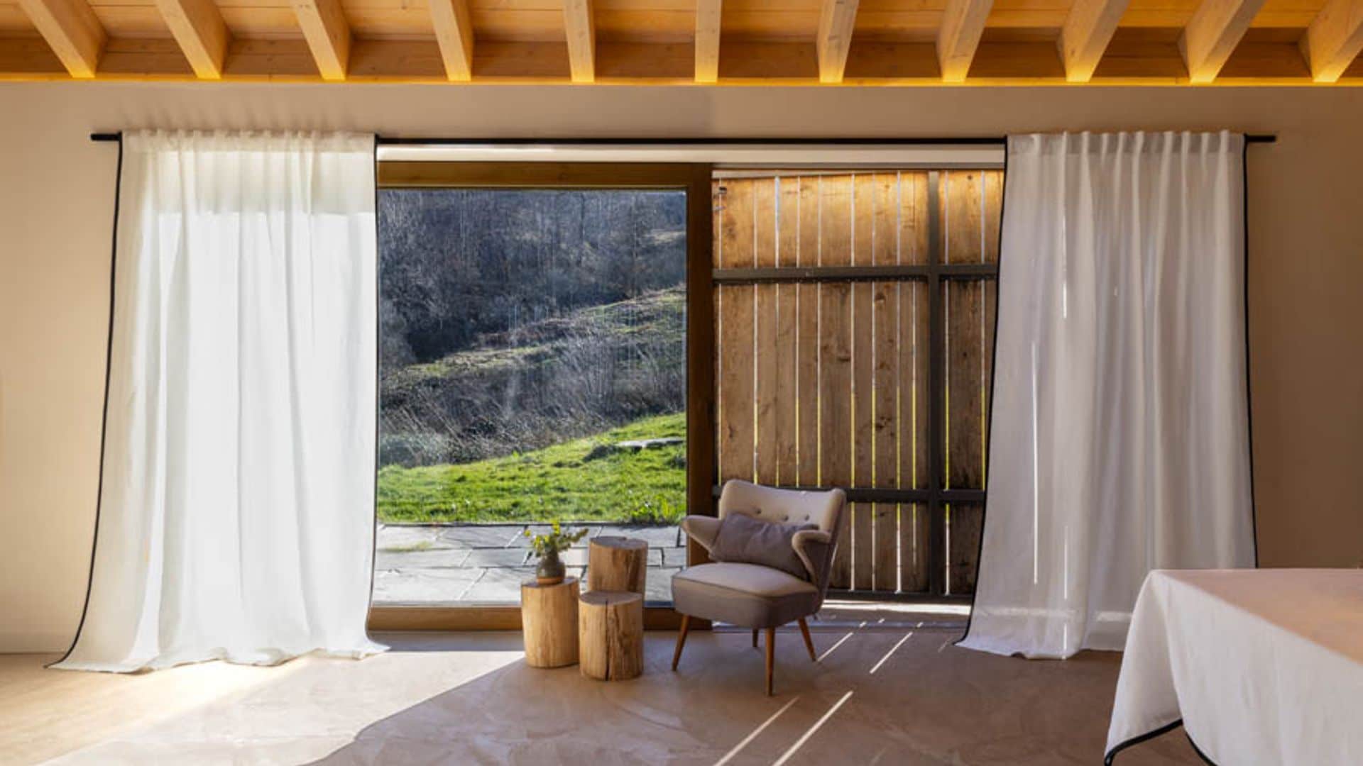 Salón con ventanas con cortinas blancas de lino, contraventanas de madera, butaca gris, mesas auxiliares tipo tronco, mesa con mantel