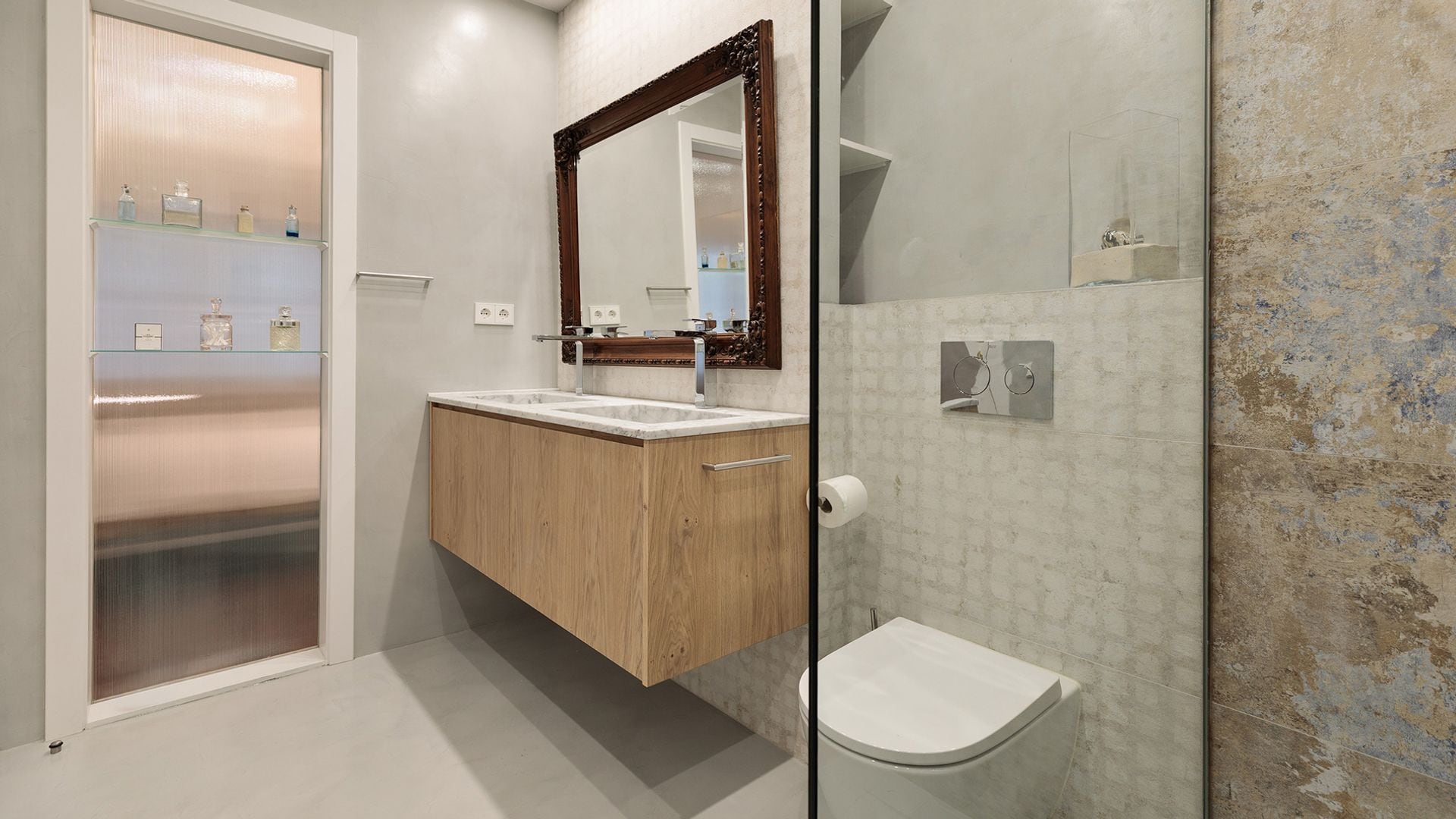 Cuarto de baño con mueble suspendido de madera, espejo con el marco de madera, baldas de cristal con botes de perfume, mampara 