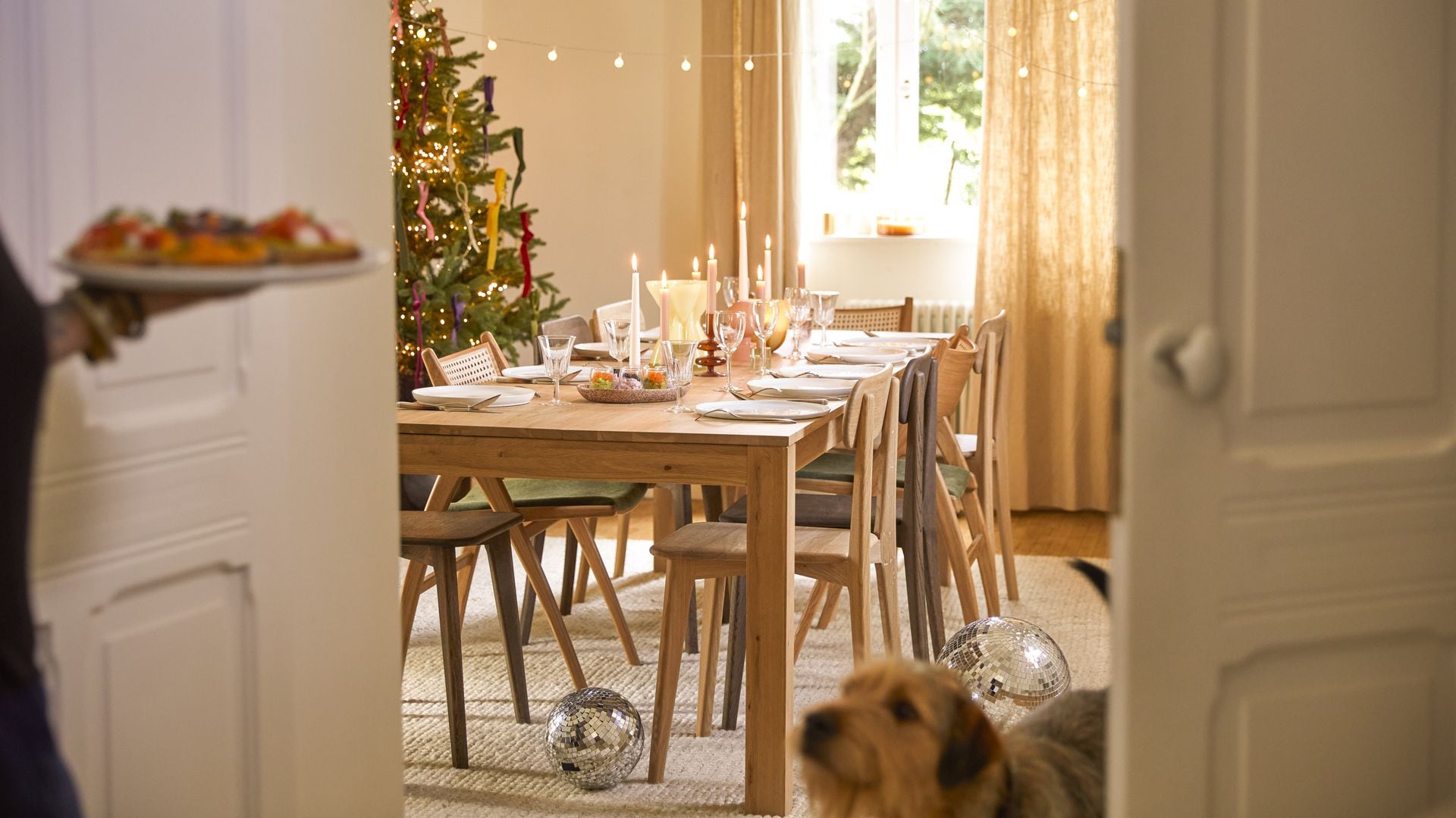 Comedor con mesa y sillas de madera, árbol de Navidad, mesa con candelabros y vajilla de fiesta, guirnalda de luces, mujer con bandeja y perro