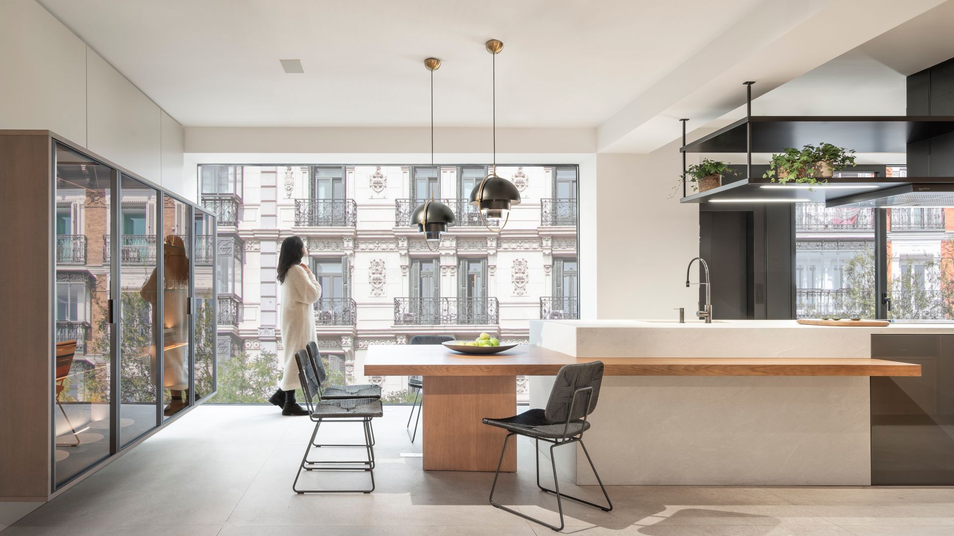 Cocina con mujer mirando por la ventana, isla, armario vitrina de pared a pared, encimera de madera, campana decorativa y lámparas de techo negras 