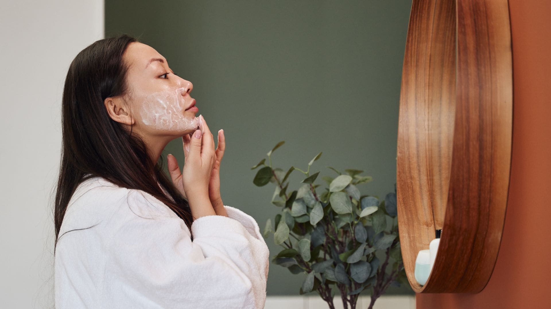 Mujer joven asiática aplicándose crema en el rostro frente al espejo de su cuarto de baño