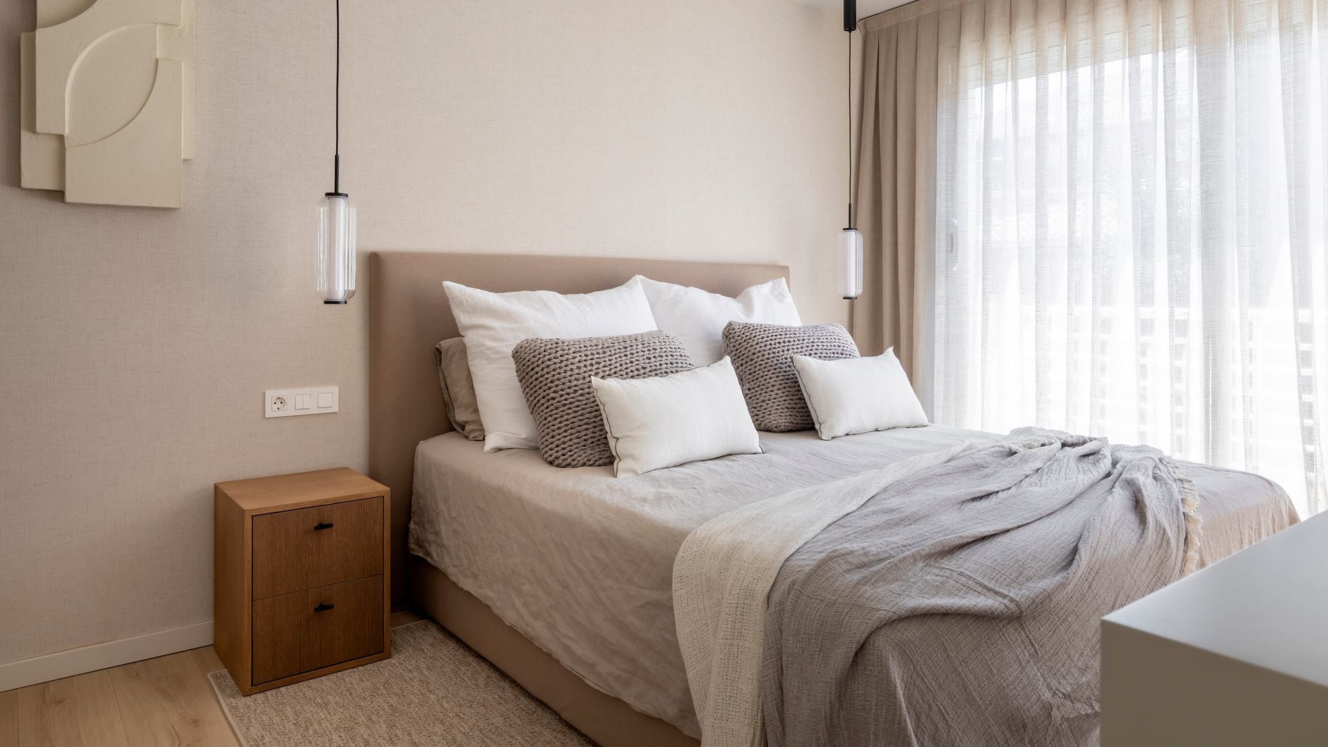 Dormitorio en tonos tierra con ropa de cama de lino y algodón, cortinas blancas, paredes en color arena, alfombra, mesita de noche, lámparas de techo a los lados de la cama, cabecero tapizado