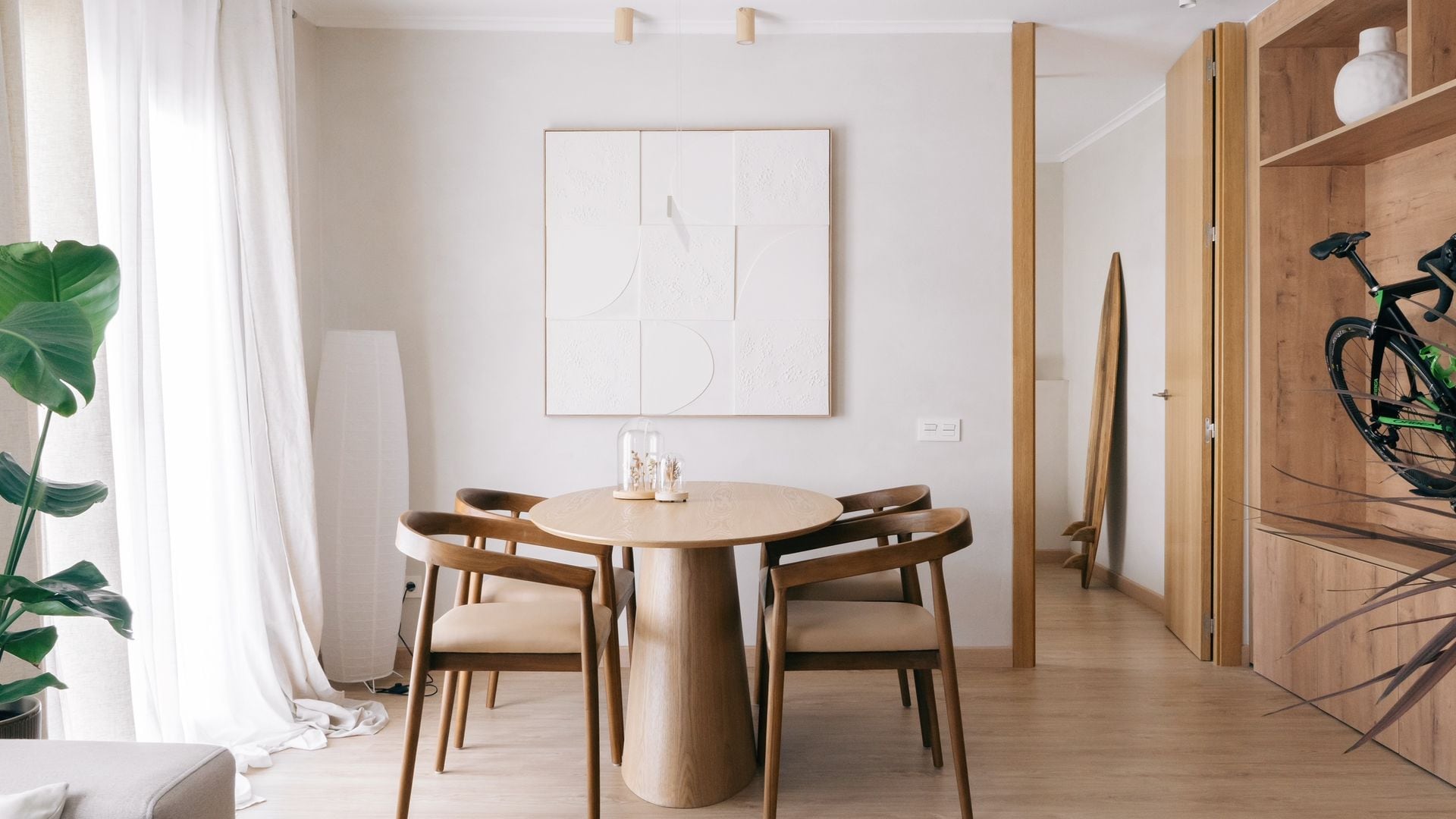 Comedor con mesa ovalada de madera, sillas de madera con el asiento tapizado, cortinas blancas, suelo de madera, cuadro en la pared