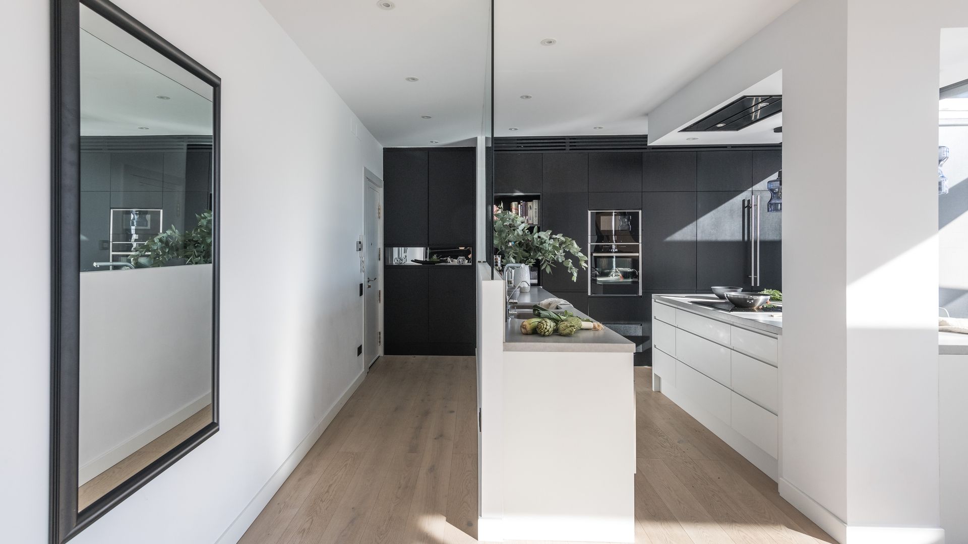 Cocina blanca y gris, suelo de madera, separada de la entrada por un medio tabique de cristal, espejo con marco negro y suelo de madera