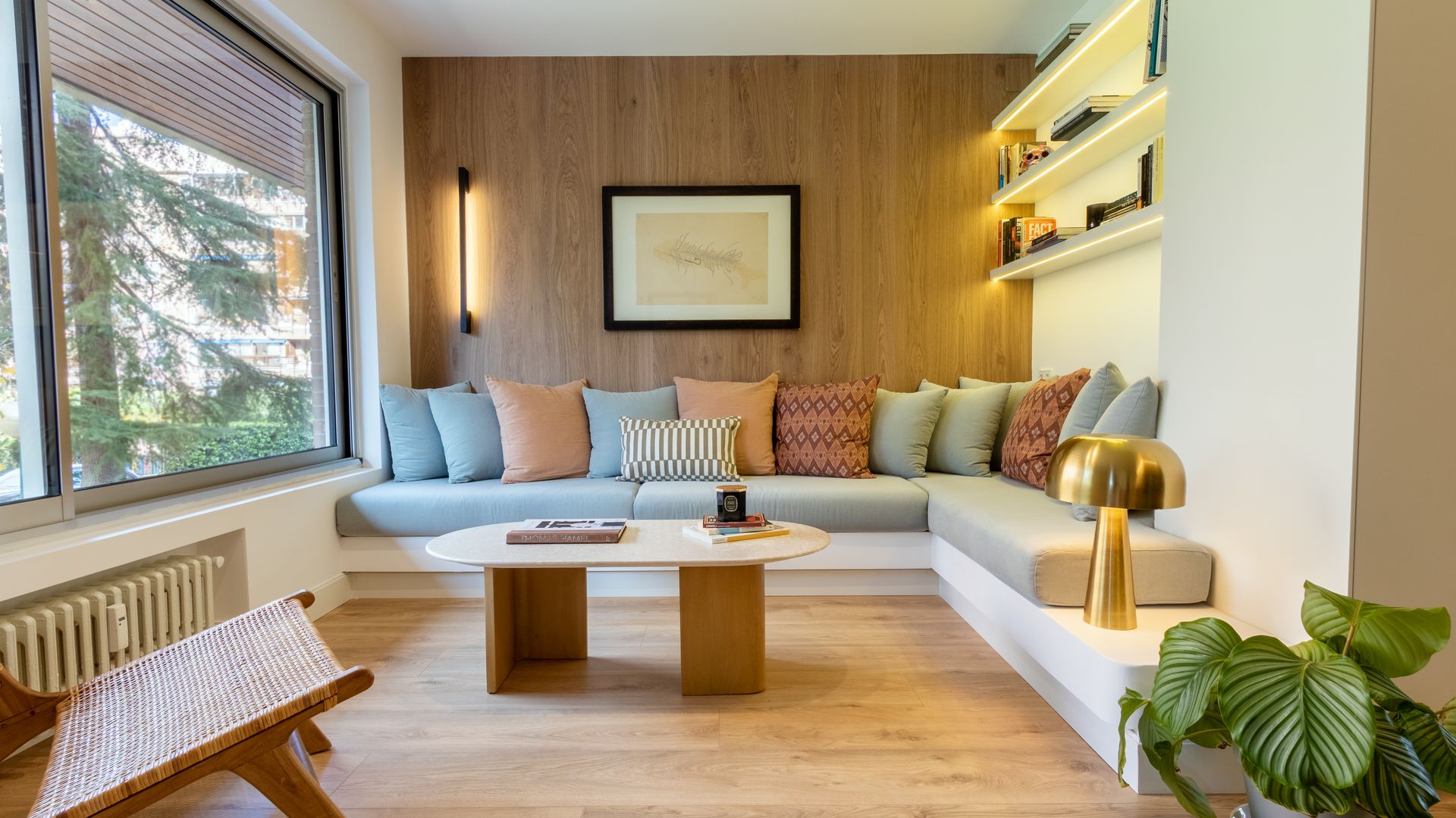 Salón con librería de obra, sofá de obra tipo banco con almohadones, mesa de centro de madera, apliques de pared, gran ventanal 