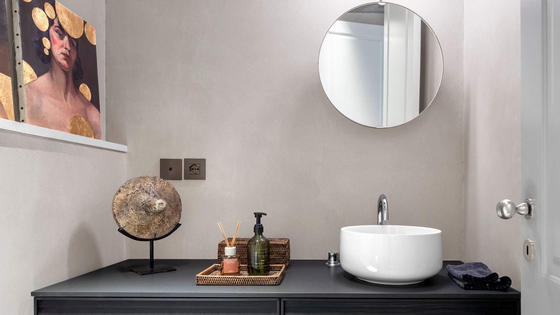Baño con mueble bajolavabo negro, espejo redondo, cuadros, lavabo blanco mini, bandeja con jabón y mikado