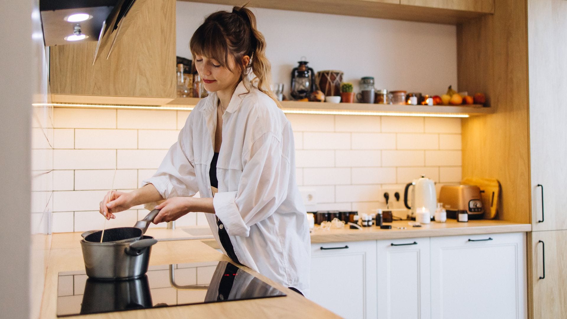 Mujer cocinando en la placa de inducción de una cocina en madera y blanco. Está moviendo un cazo al fuego.
